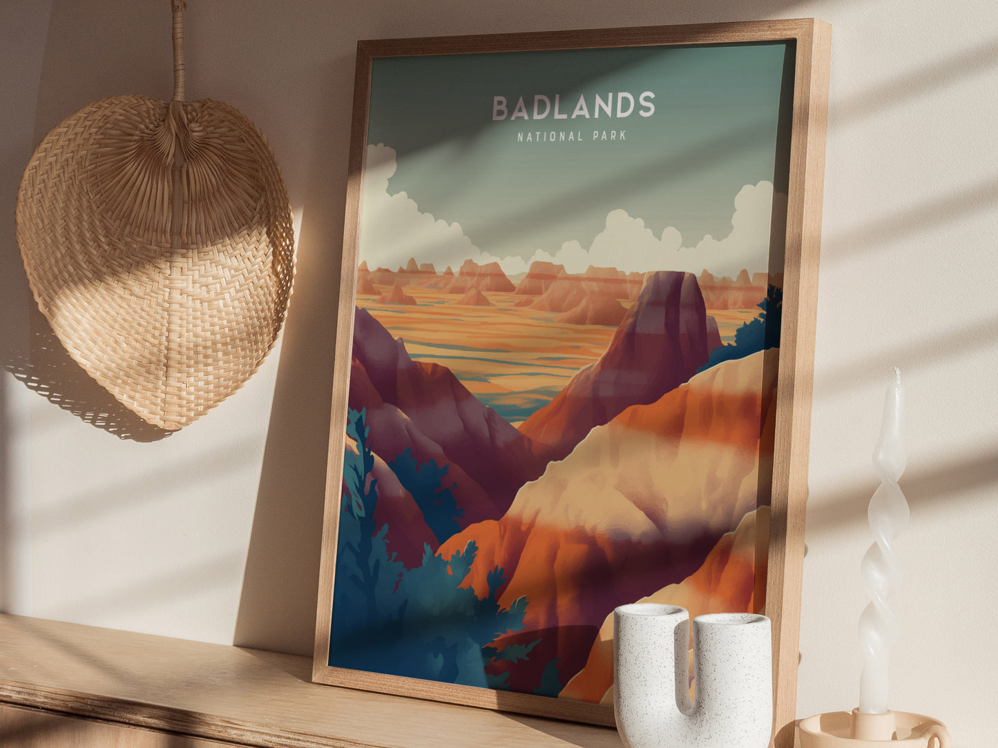 A framed Badlands National Park poster showcasing colorful rock formations is displayed on a wooden shelf, alongside a woven basket, a white ceramic vase, and a twisted candle.