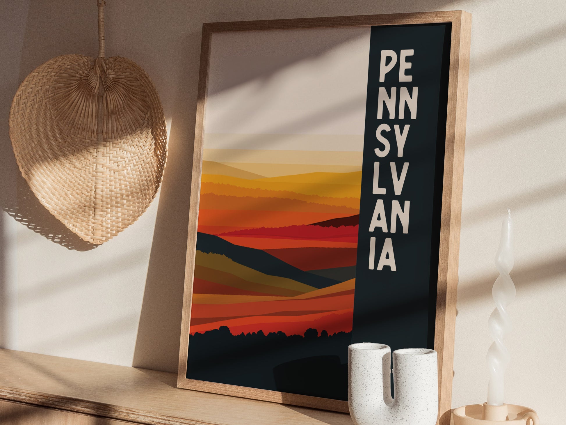 A framed abstract art print of Pennsylvania with bold colors and vertical text, placed on a wooden shelf alongside a woven wall decoration and modern ceramic vase.