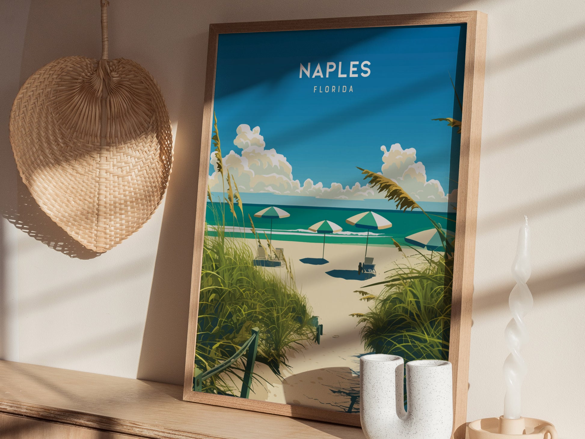 A framed poster of a beach scene with umbrellas is displayed on a shelf, accompanied by a woven fan, a candle, and decorative vases. Sunlight casts soft shadows.