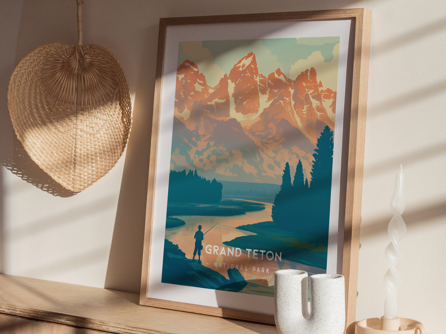 A framed minimalist poster of Grand Teton National Park is displayed on a wooden shelf, alongside a woven basket and a twisted white candle. Sunlight casts shadows across the scene.