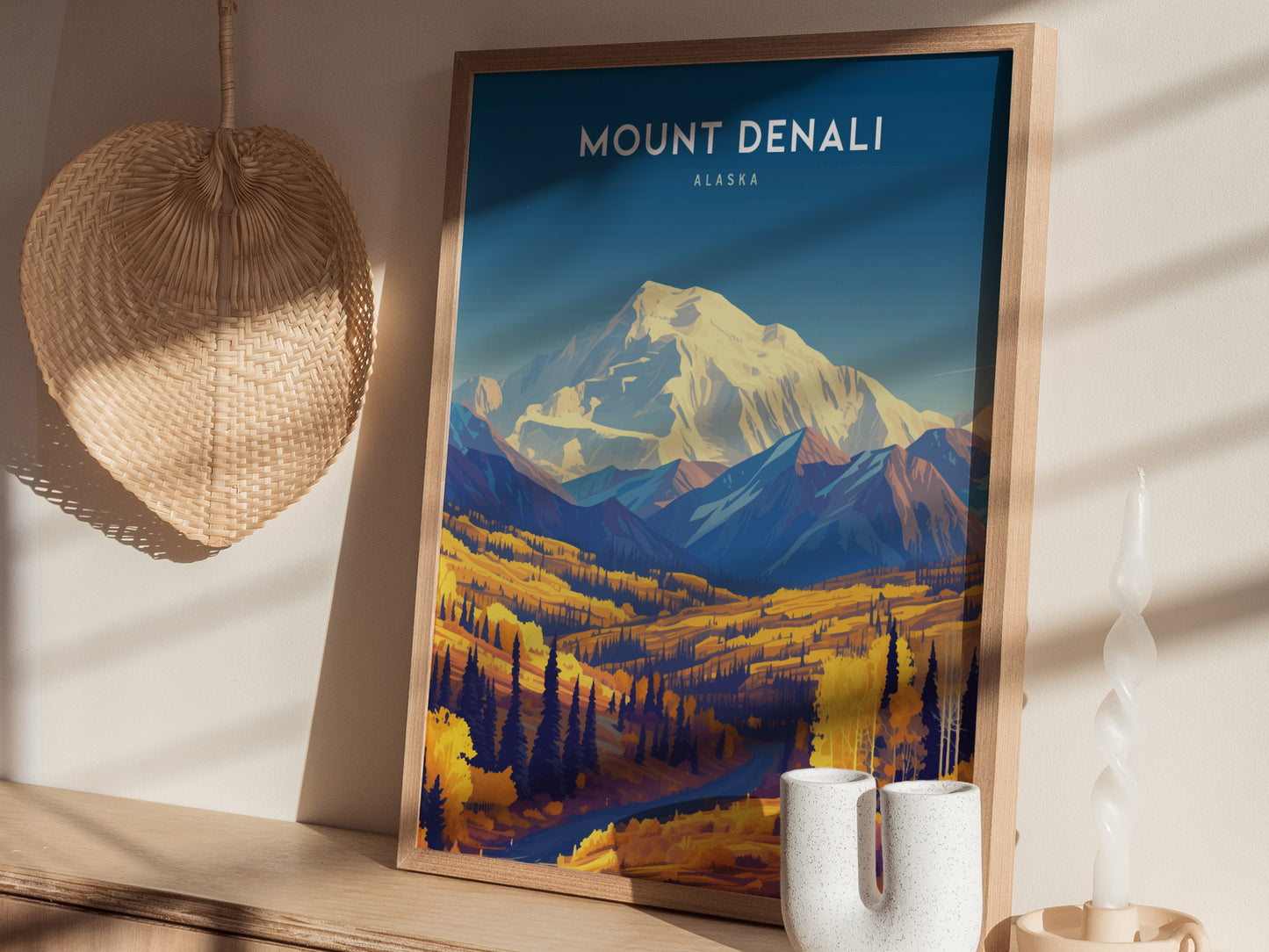 A framed Mount Denali poster with a vibrant landscape rests on a wooden shelf, accompanied by a woven basket and a unique white candle.