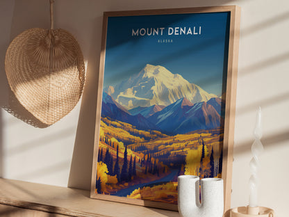A framed Mount Denali poster with a vibrant landscape rests on a wooden shelf, accompanied by a woven basket and a unique white candle.