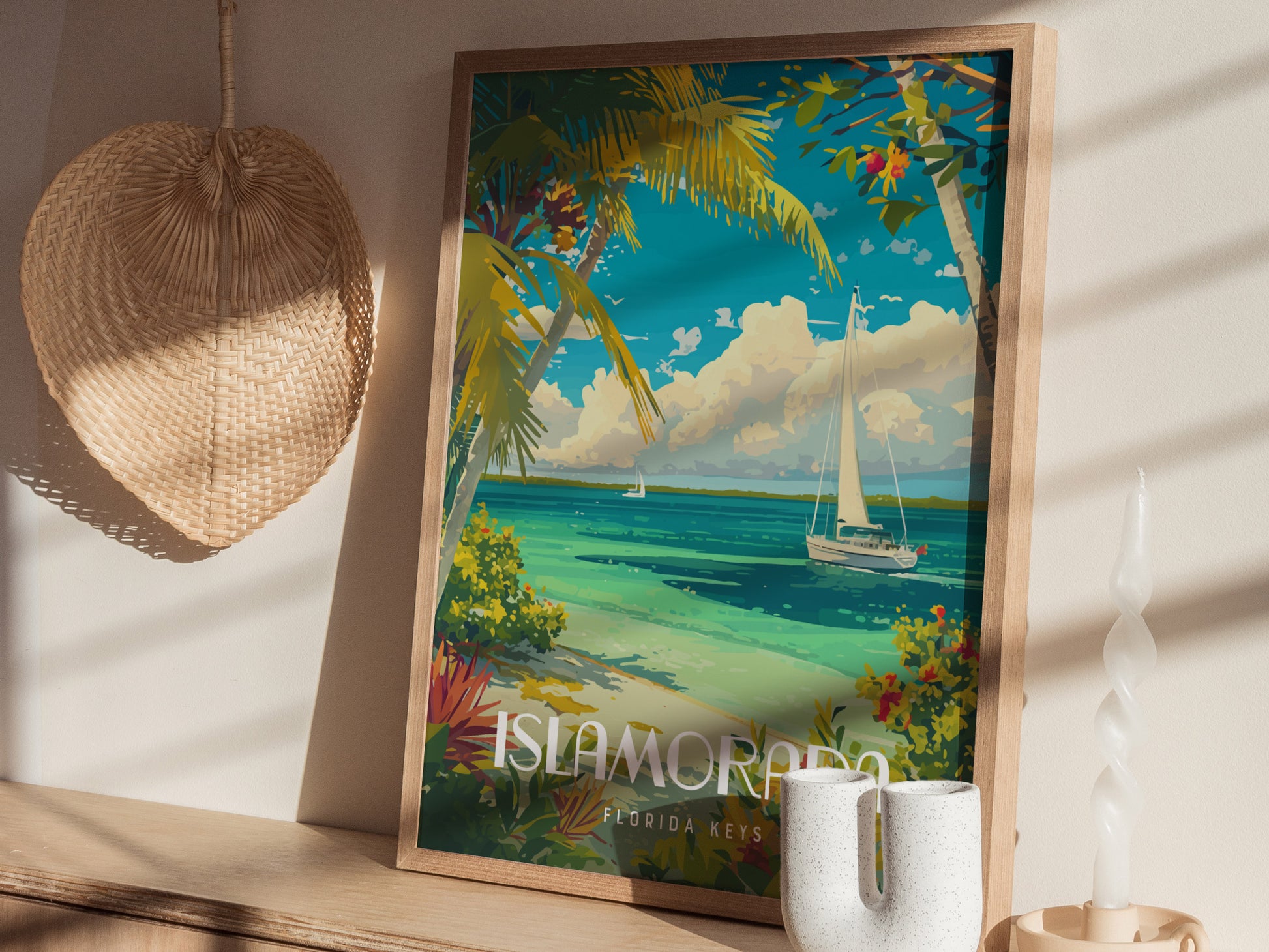 A framed Islamorada poster shows a vibrant coastal scene with turquoise water, sailboat, and palm trees. Its displayed on a wooden shelf beside a woven fan and candle.