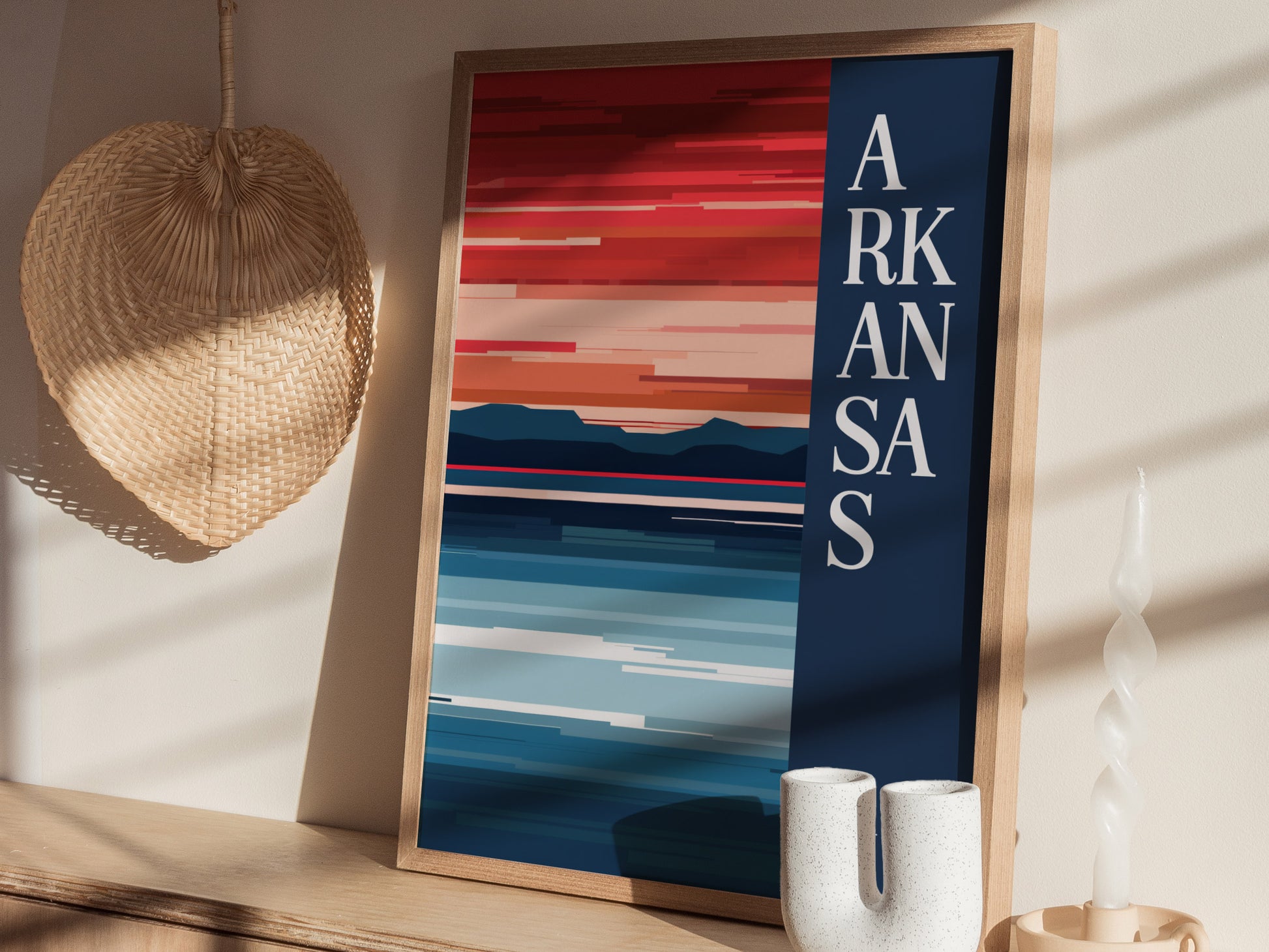 A framed Arkansas minimalist design poster features abstract landscapes in bold colors with the word Arkansas vertically. It sits on a wooden shelf alongside a wicker fan and decorative items.