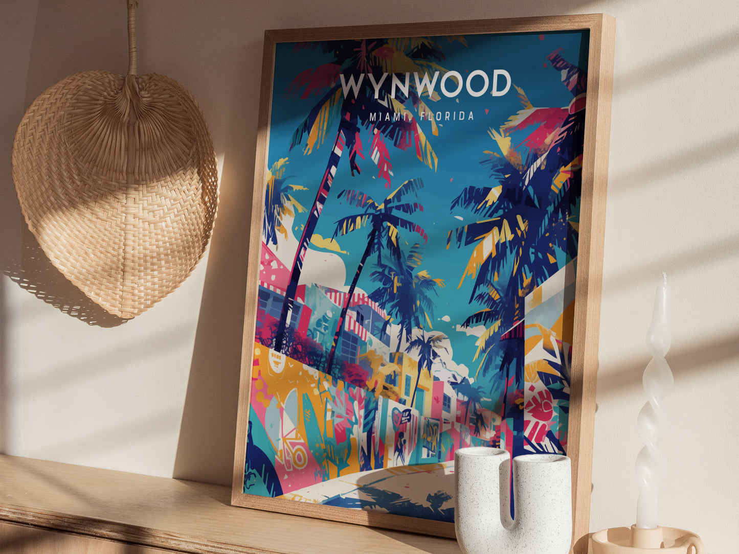 A colorful poster of Wynwood, Miami, featuring vibrant graffiti murals and palm trees, is displayed in a wooden frame. It rests on a wooden surface alongside a woven fan and a uniquely shaped white candle.