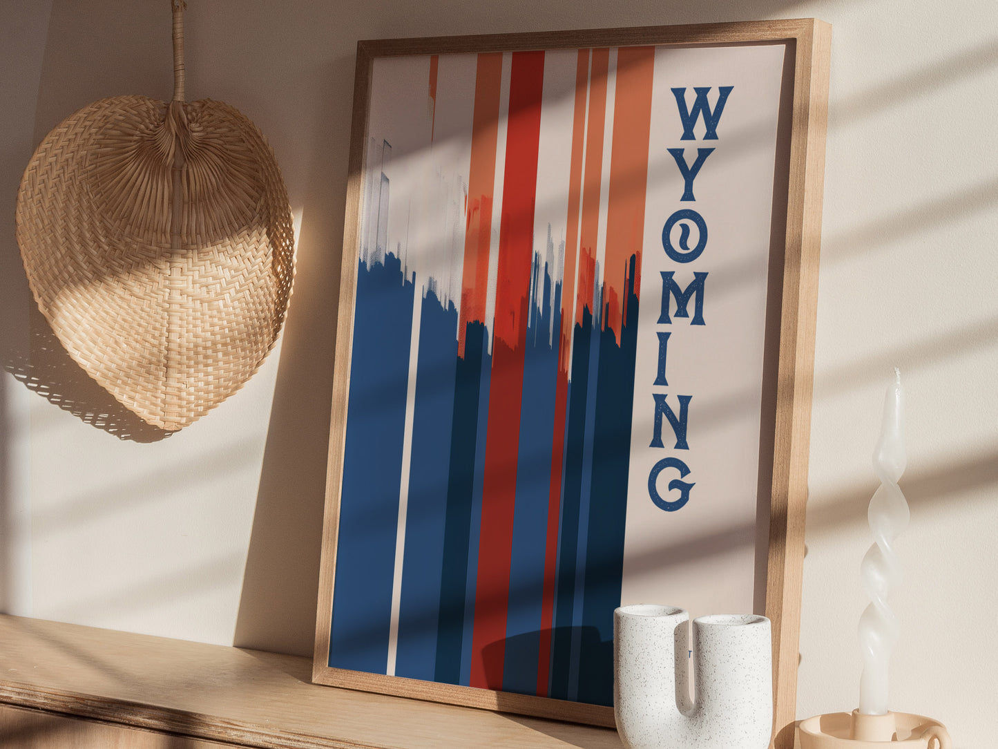 A framed Wyoming poster with bold red, white, and blue vertical stripes and text stands on a shelf near a woven hat and a twisted white candle.