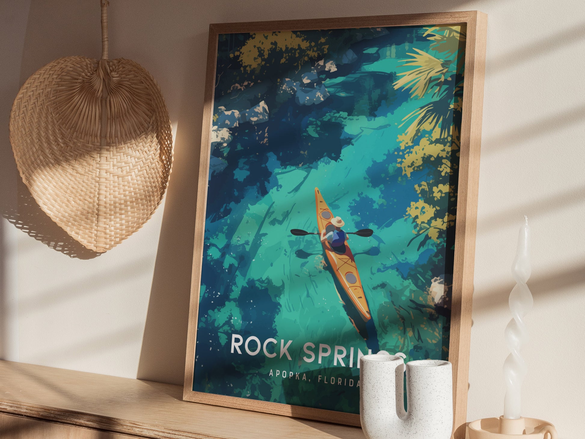 A framed poster of a kayaker on clear turquoise water is displayed on a wooden surface, accompanied by a wicker fan and a twisted white candle.