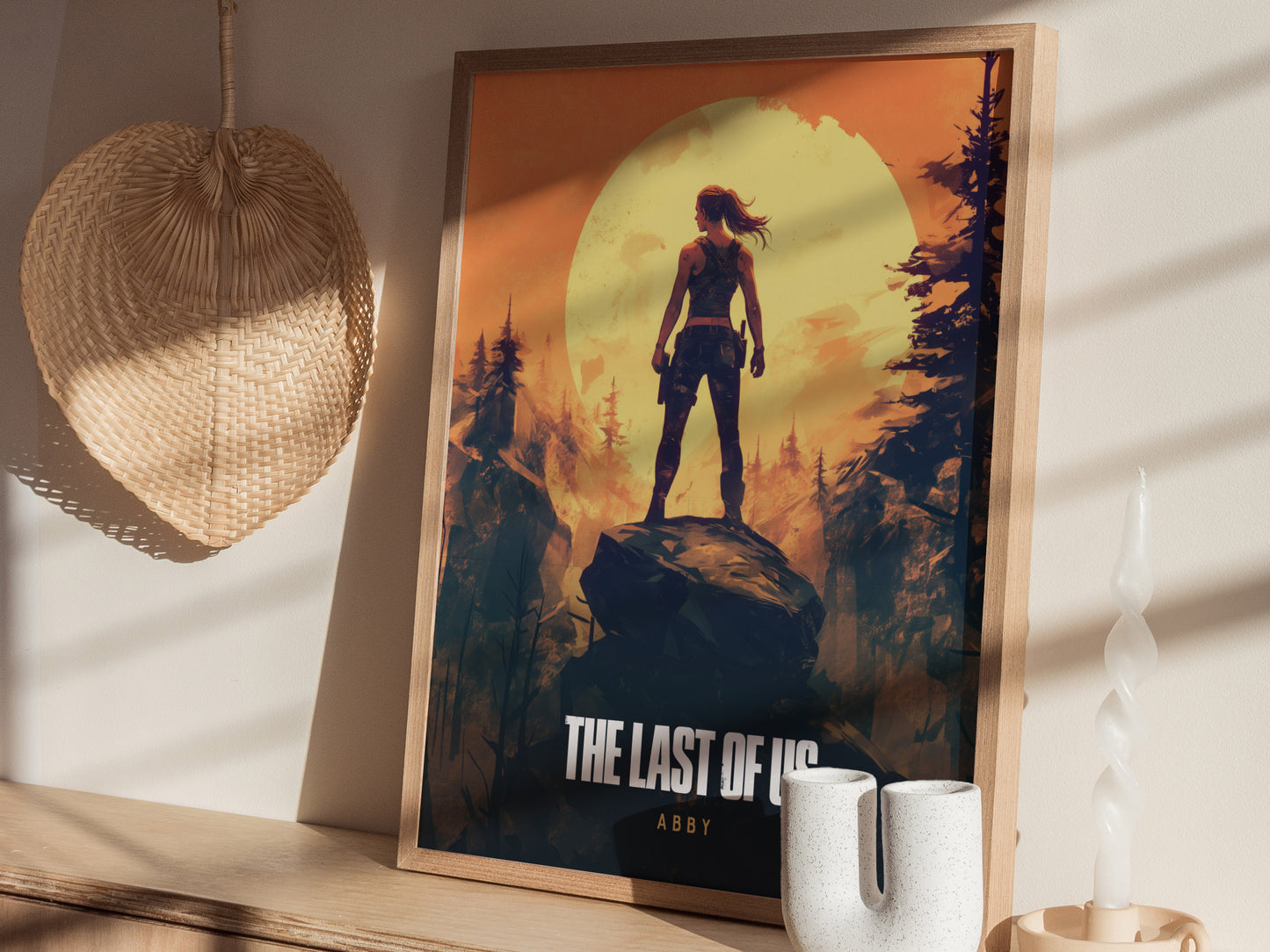 Framed poster featuring a silhouetted figure standing on a rock against a large yellow sun and forest background. Its displayed on a wooden surface alongside a woven basket and candle.