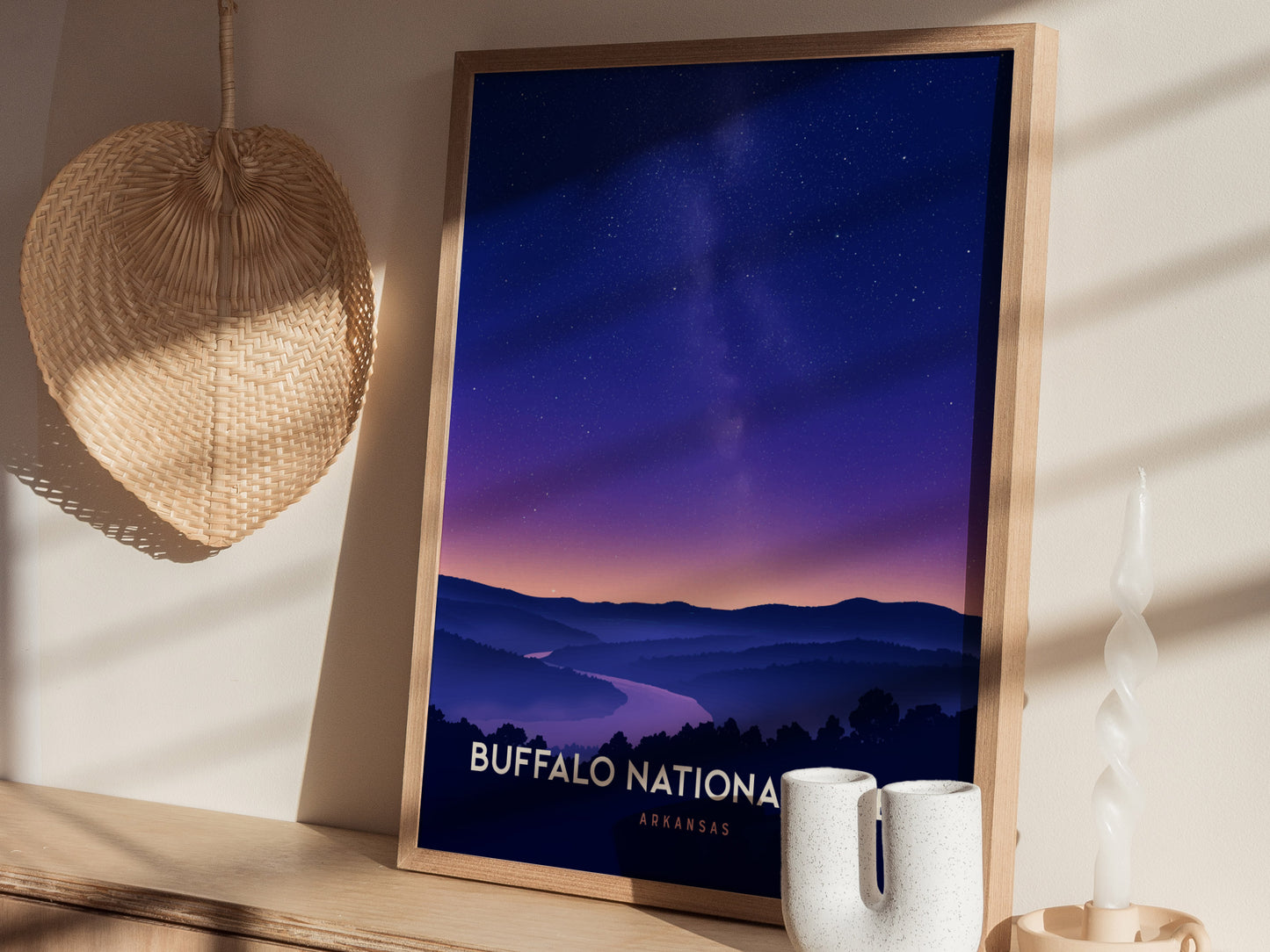 A framed travel print titled Buffalo National is displayed on a shelf, showcasing a serene landscape of rolling hills and a river under a twilight sky filled with stars.