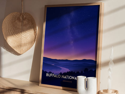 A framed travel print titled Buffalo National is displayed on a shelf, showcasing a serene landscape of rolling hills and a river under a twilight sky filled with stars.