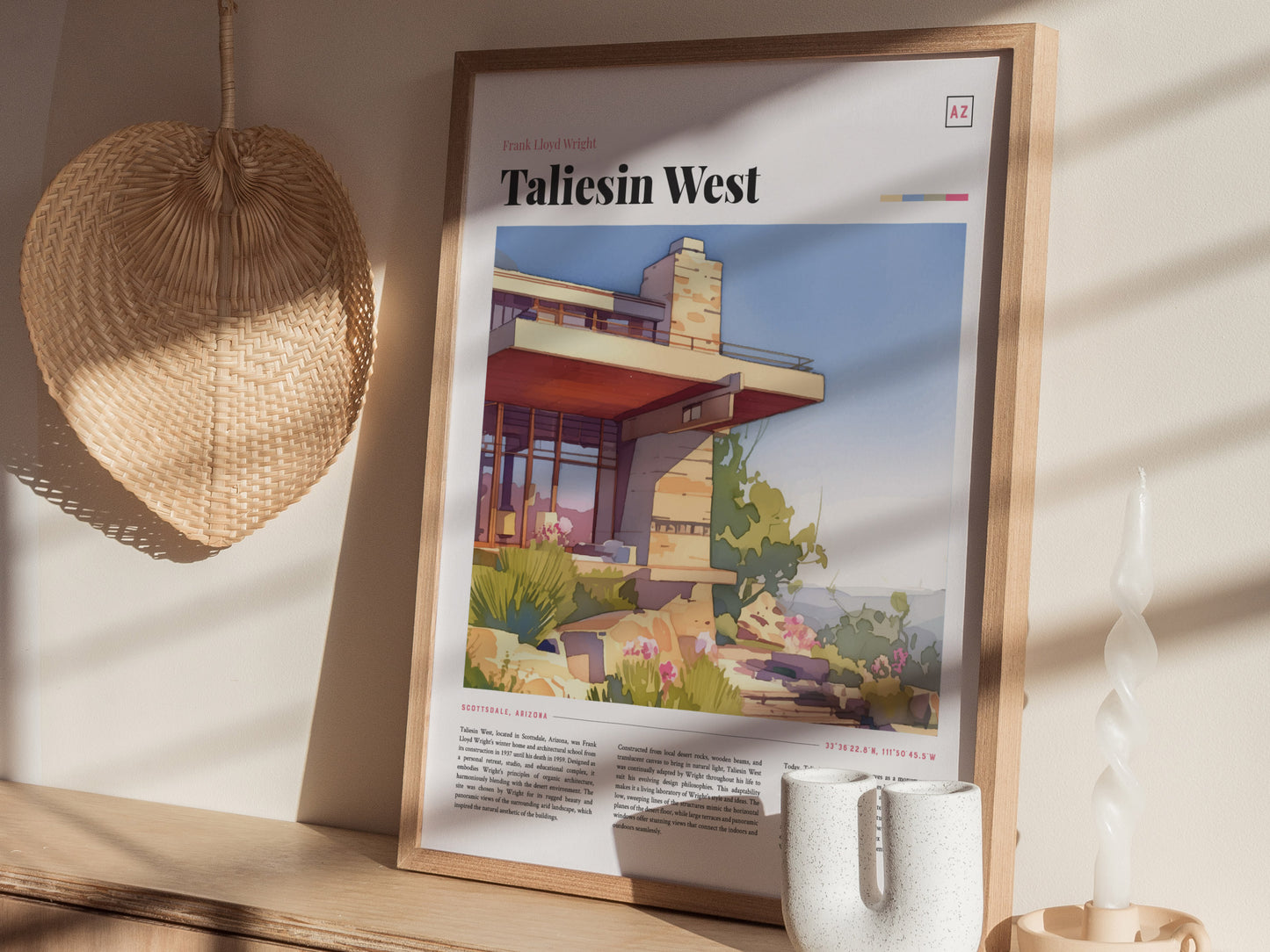 A framed Taliesin West poster is displayed on a wooden shelf, accompanied by a woven fan, a white ceramic vase, and a twisted candle, all bathed in sunlight.