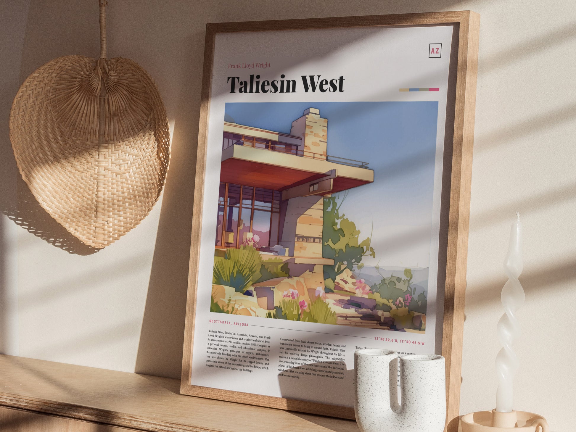 A framed Taliesin West poster is displayed on a wooden shelf, accompanied by a woven fan, a white ceramic vase, and a twisted candle, all bathed in sunlight.