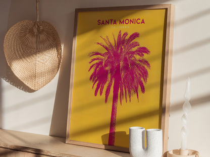 Framed poster featuring a vibrant pink palm tree against a yellow background, labeled Santa Monica, California. Displayed next to a woven fan, ceramic vase, and spiral candle.