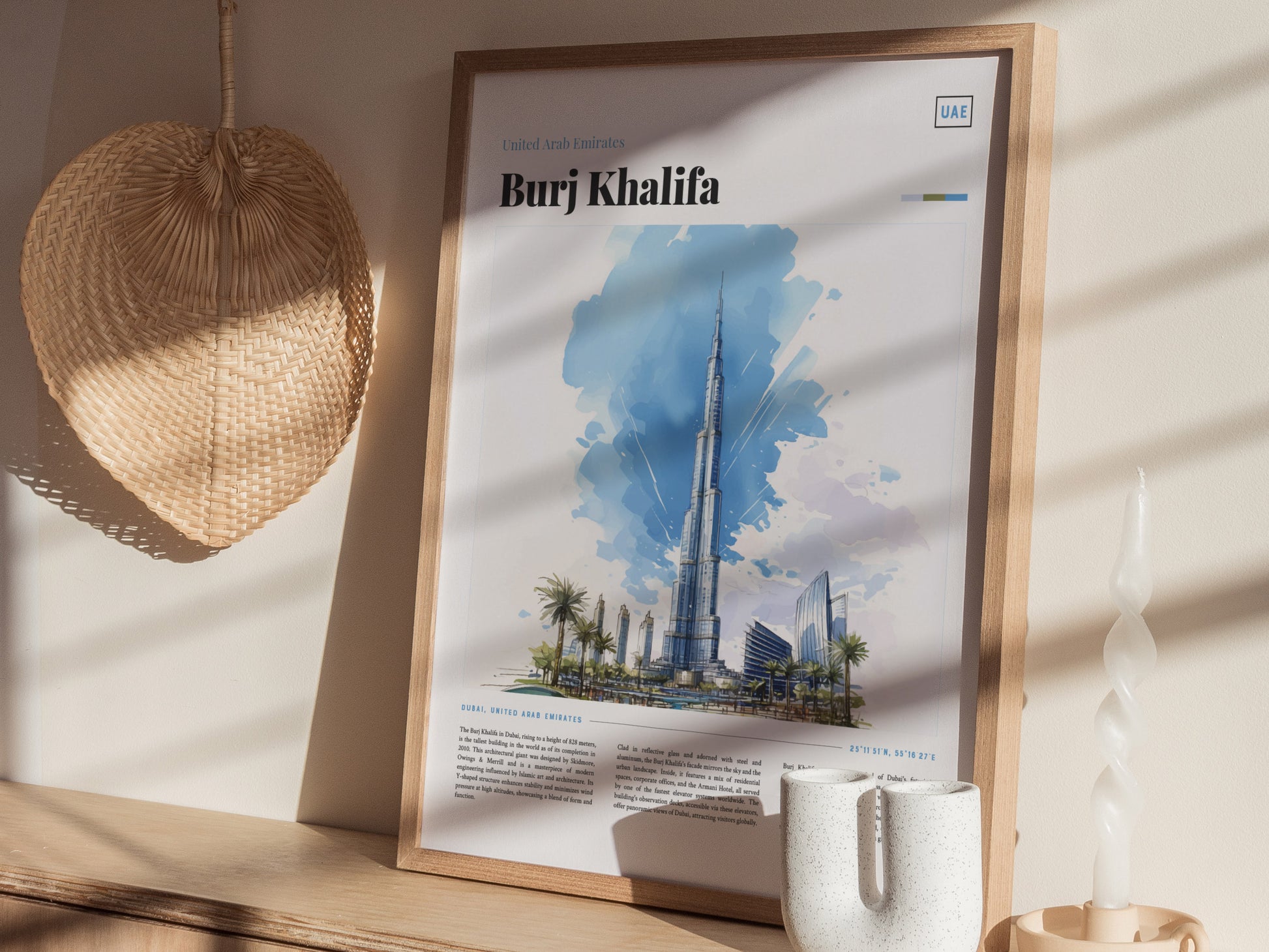 A framed poster of the Burj Khalifa with a watercolor illustration is displayed on a wooden shelf, next to a woven decorative piece, a white twisted candle, and a mug.