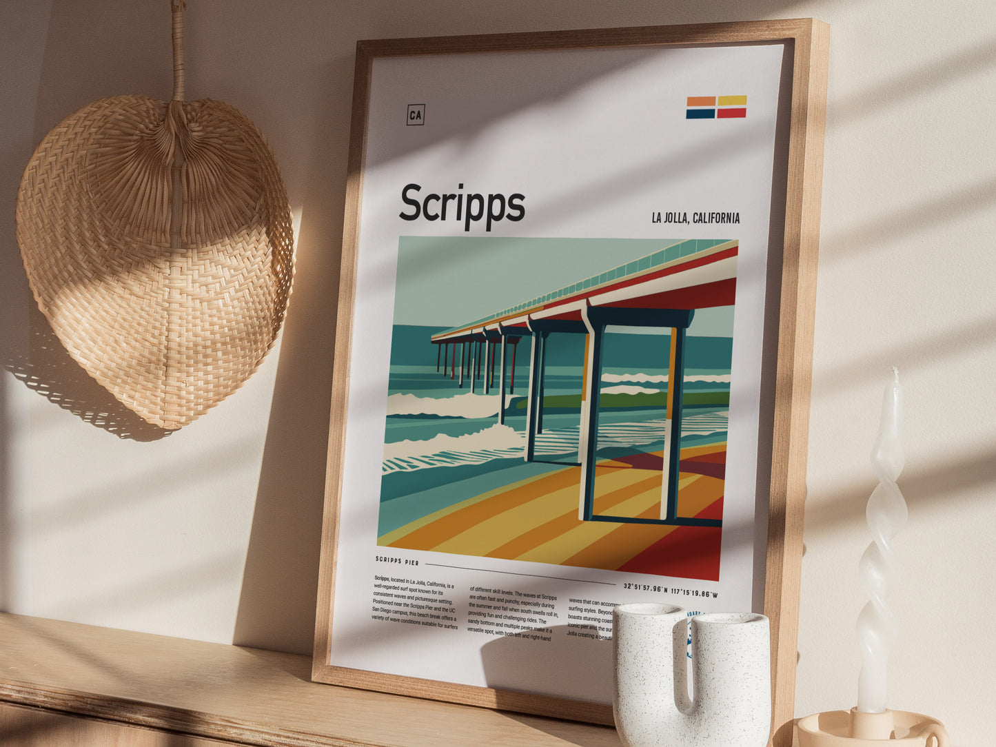 A framed poster depicting Scripps Pier with vibrant ocean waves and colorful hues is displayed on a wooden shelf, alongside a woven fan and a twisted white candle.