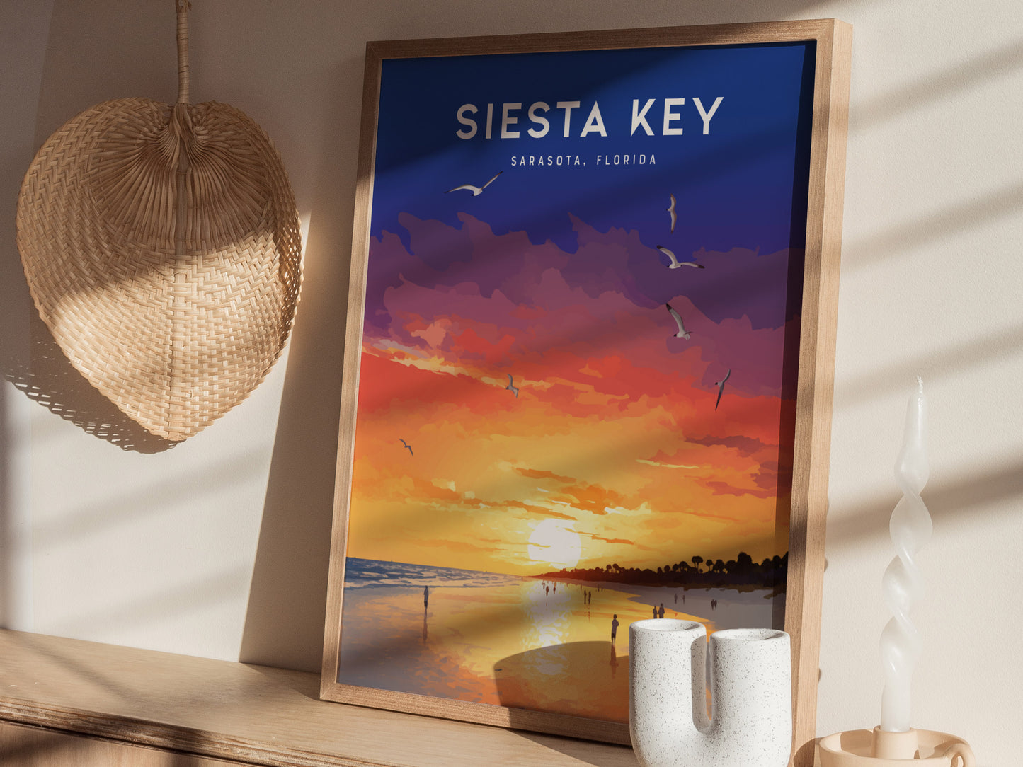 A framed poster of a vibrant Siesta Key sunset with purple, orange, and yellow hues, featuring seagulls in the sky, is displayed on a wooden shelf next to a woven.