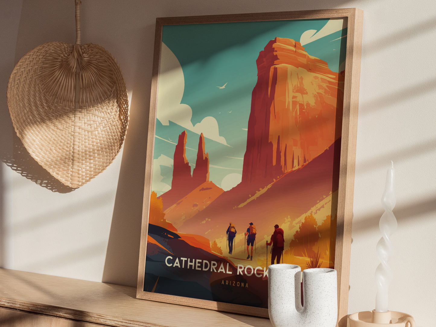A framed poster of Cathedral Rock Trail in Sedona, Arizona, featuring vivid red cliffs and hikers, is displayed on a wooden shelf next to a woven fan and candle.