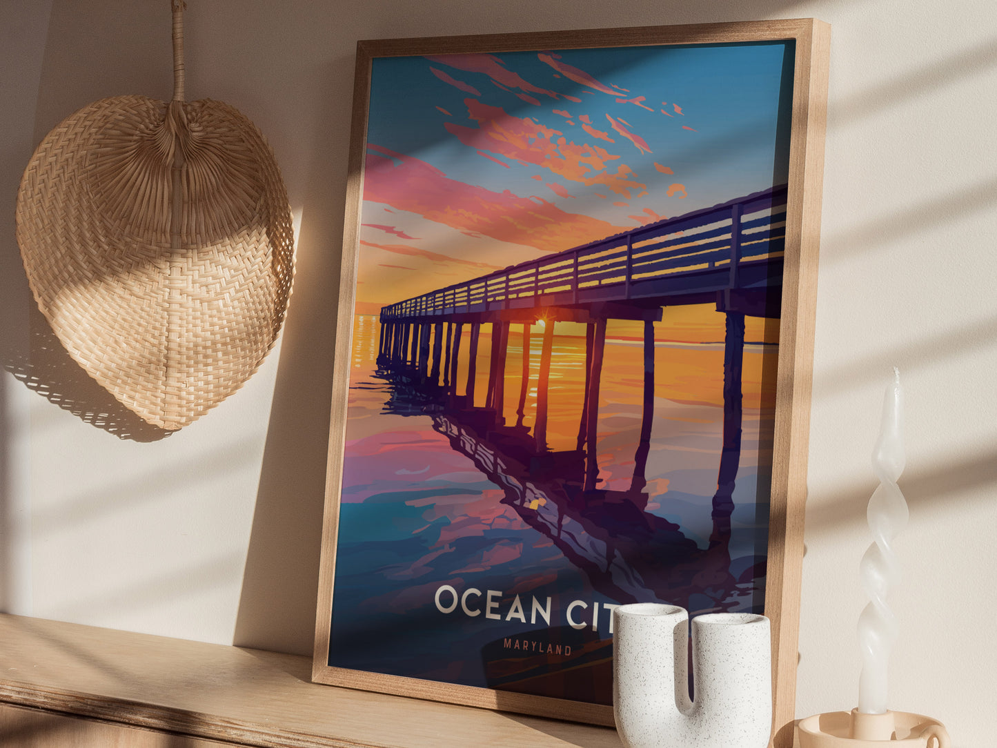 A framed poster of a sunset at a boardwalk pier is displayed on a shelf with decorative items, including a wicker fan and a twisted candle.
