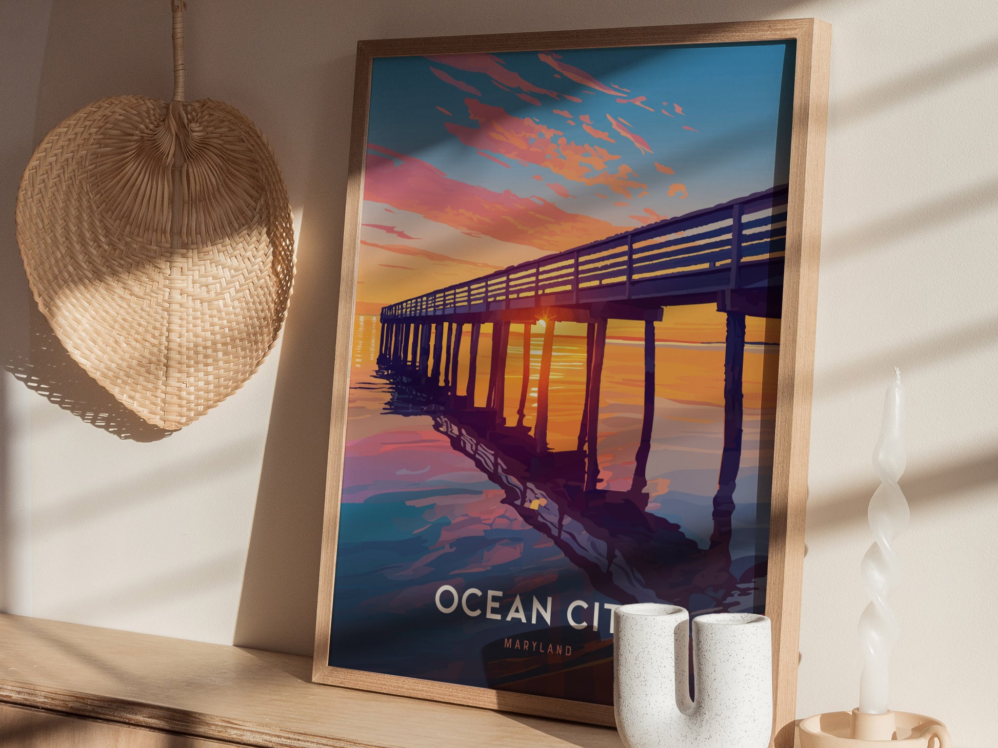 A framed poster of a sunset at a boardwalk pier is displayed on a shelf with decorative items, including a wicker fan and a twisted candle.