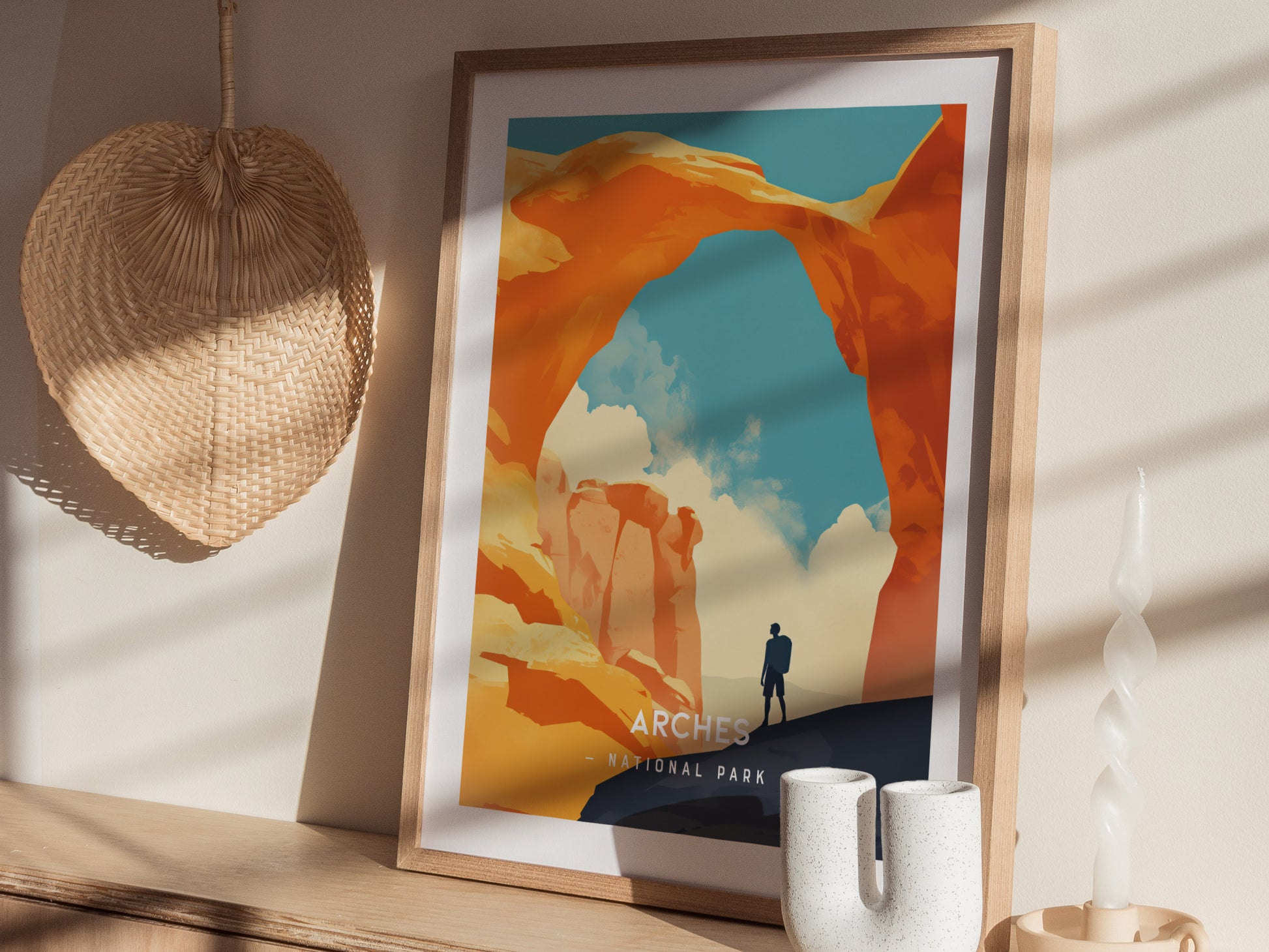 A framed minimalist poster of Arches National Park featuring Delicate Arch against a blue sky, placed on a wooden shelf next to a woven fan and decorative items.