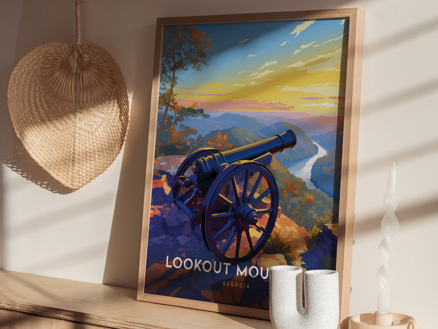 A framed poster depicting a cannon overlooking a scenic mountain landscape at sunset, set against a wall with a woven basket and decorative items nearby.