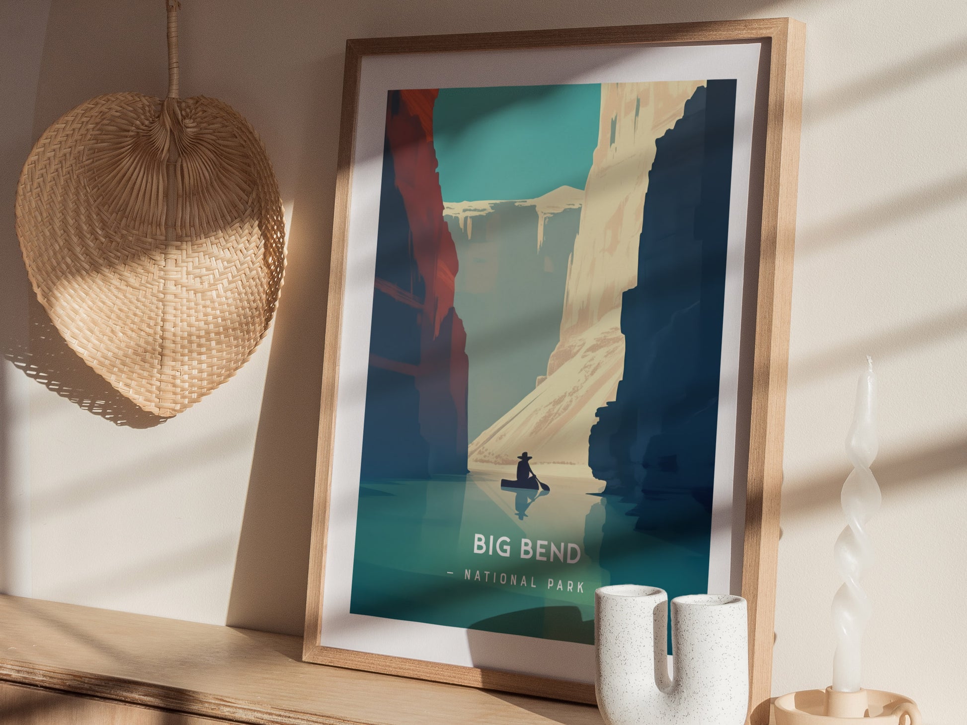 A framed poster depicting Big Bend National Park is displayed on a wooden shelf, alongside a woven fan, a ceramic vase, and a twisted white candle.