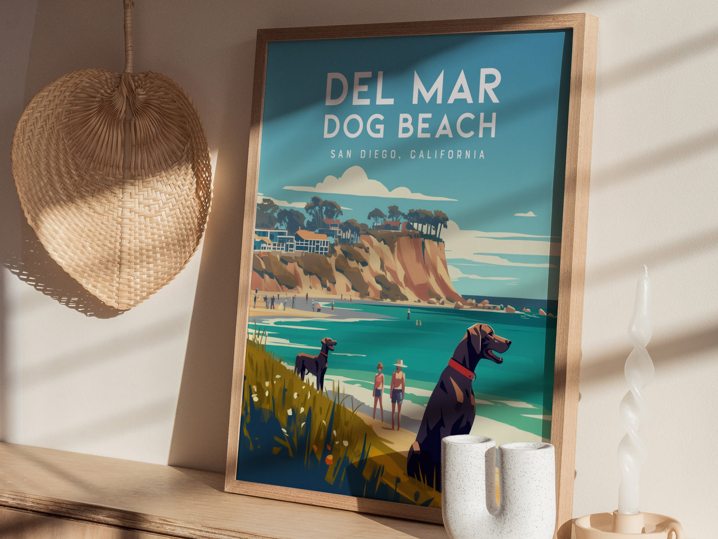 Framed poster of Del Mar Dog Beach with vibrant coastal illustration featuring dogs, people, and cliffs. Displayed on a wooden shelf with decorative items like a woven fan and candle.