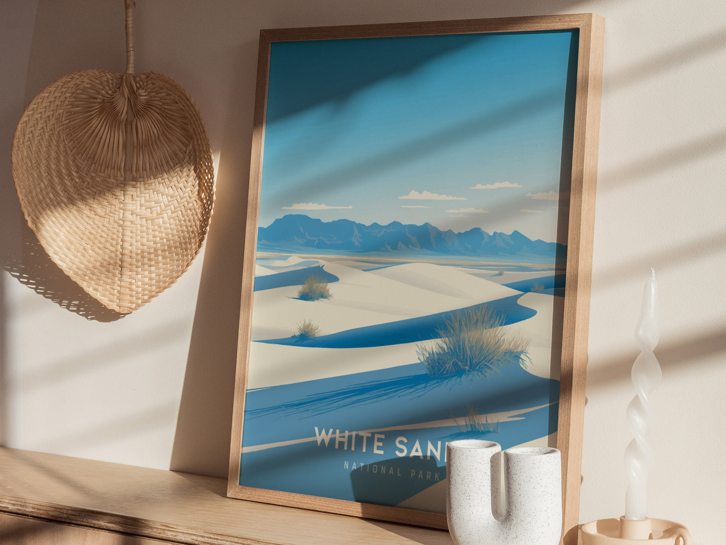 A framed poster depicting White Sands National Park with blue skies, white dunes, and distant mountains, is displayed on a shelf alongside a woven wall decor and a twisted candle.