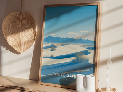 A framed poster depicting White Sands National Park with blue skies, white dunes, and distant mountains, is displayed on a shelf alongside a woven wall decor and a twisted candle.