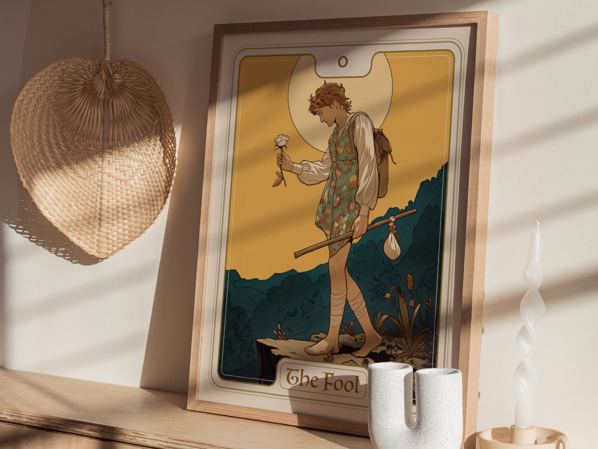 A framed poster of The Fool tarot card is displayed on a wooden shelf with a woven fan and decorative candle nearby, casting soft shadows.