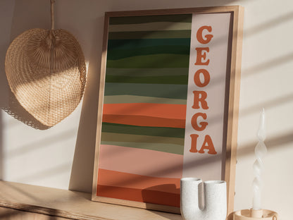 Framed Georgia poster with bold typography and earthy horizontal stripes rests on a wooden shelf, beside a twisted candle and a woven wall decoration. Sunlight casts gentle shadows.