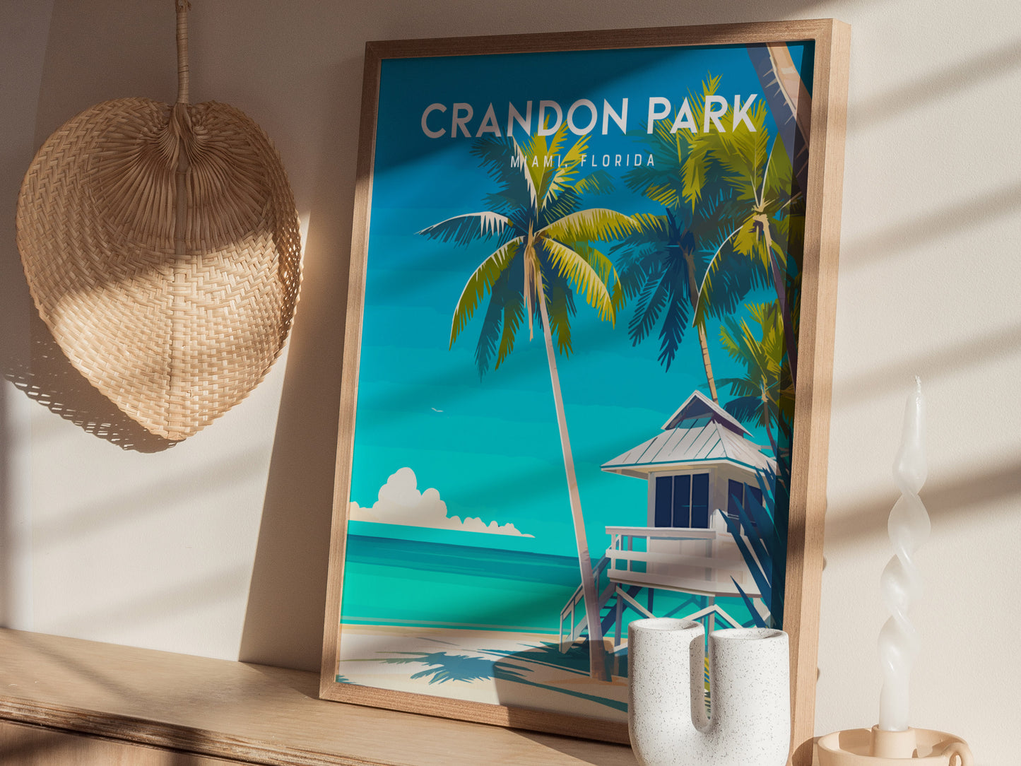 A framed poster of Crandon Park Beach, Miami, depicts palm trees and a lifeguard tower against turquoise waters. It leans against a wall with a woven basket and a sculpted candle nearby.