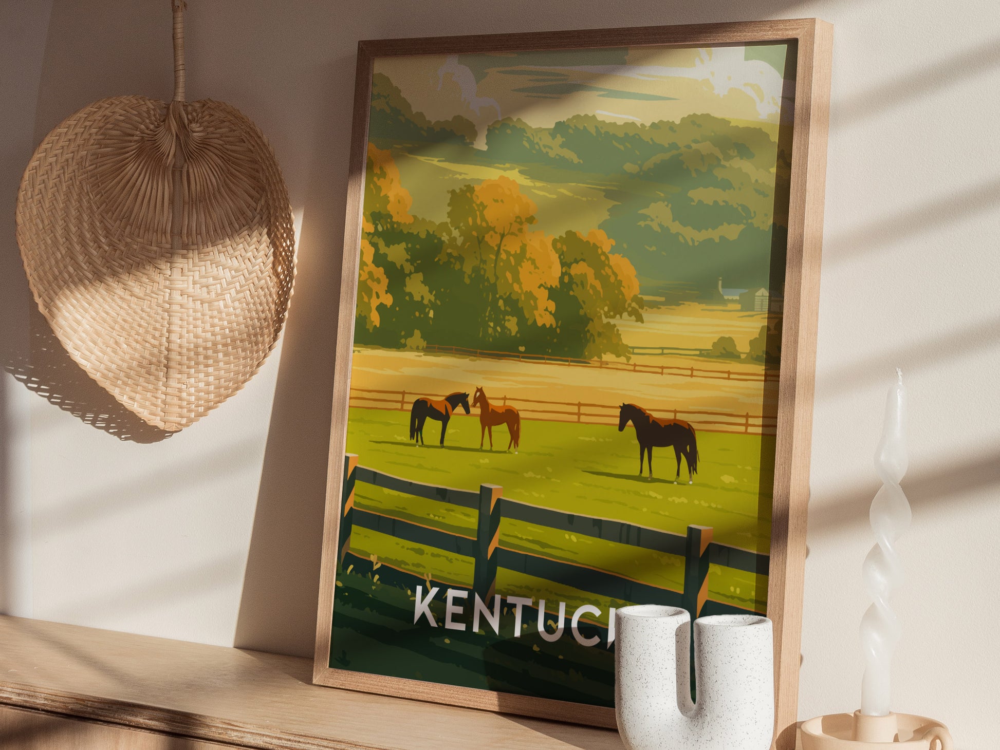 A framed poster depicting a Kentucky landscape with horses grazing in a green pasture, set against a wooden table with a woven hat and decorative candle nearby.