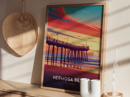 A framed poster of Hermosa Beach Pier at sunset is displayed on a wooden surface, surrounded by a woven basket and a twisted white candle.