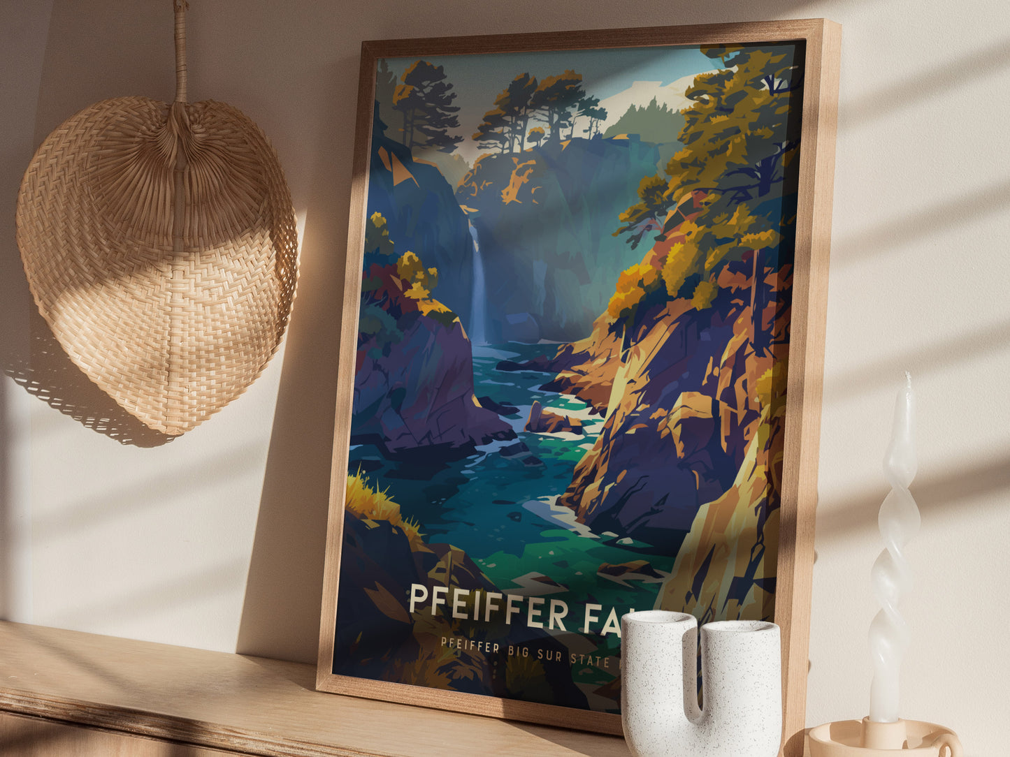 A framed poster of Pfeiffer Falls in Big Sur, featuring a colorful landscape with trees and a waterfall, is displayed on a wooden surface alongside a woven basket and decorative.