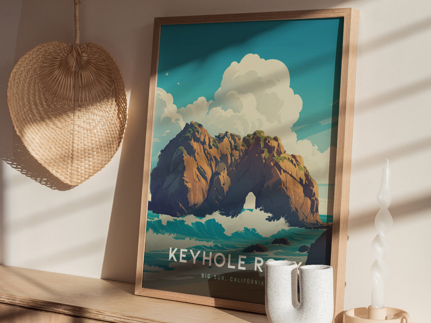 The image shows a framed poster of Keyhole Rock at Pfeiffer Beach, featuring a dramatic seascape with waves and clouds. It rests on a wooden surface next to a woven basket and a uniquely shaped white candle.