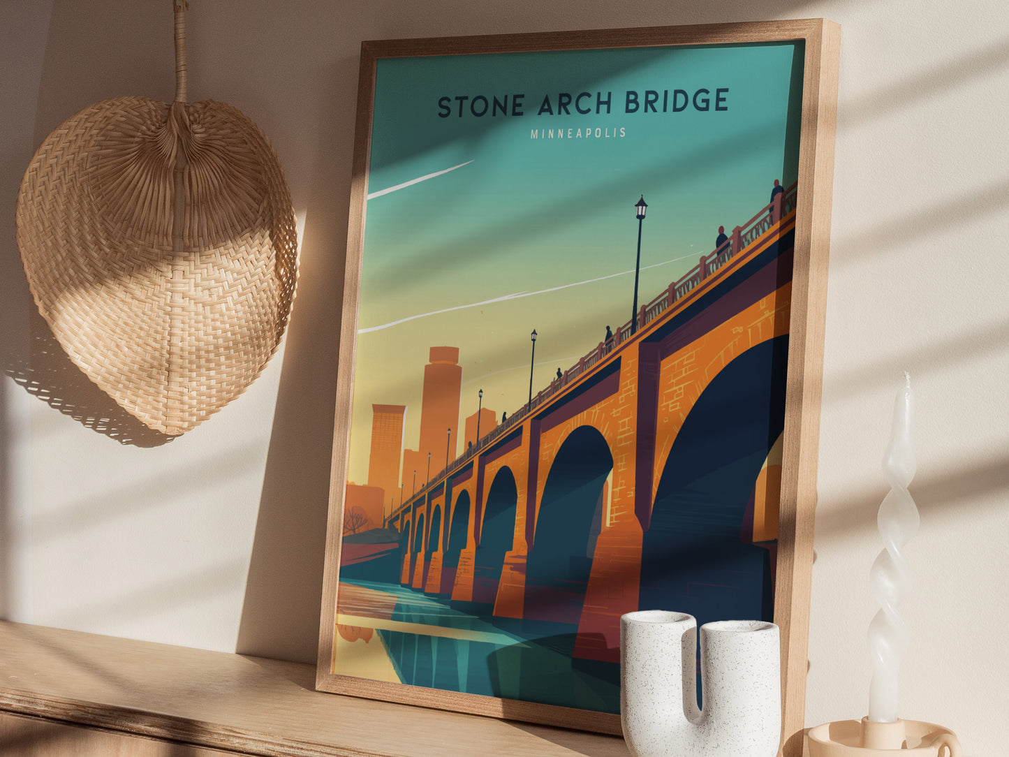 A framed poster of the Stone Arch Bridge in Minneapolis is displayed on a wooden shelf. Nearby are a woven straw wall hanging and a white twisted candle.