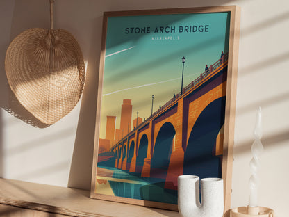A framed poster of the Stone Arch Bridge in Minneapolis is displayed on a wooden shelf. Nearby are a woven straw wall hanging and a white twisted candle.