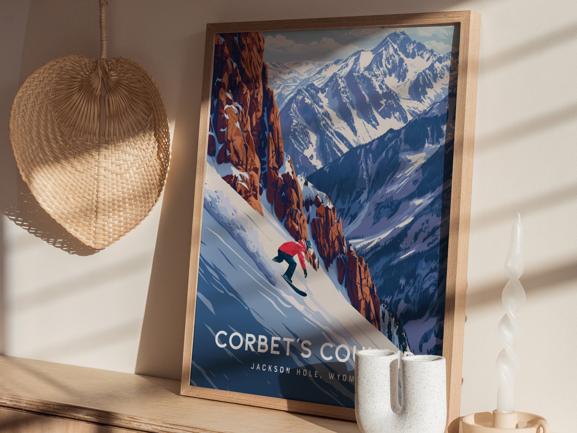A framed poster of a snowboarder descending a snowy mountain with rugged peaks in the background, displayed on a wooden shelf with a woven fan and decorative candle.