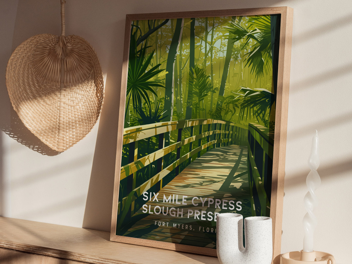 A framed poster of Six Mile Cypress Slough Preserve shows a wooden boardwalk through lush greenery. It is displayed on a shelf with a woven fan and decorative candle.