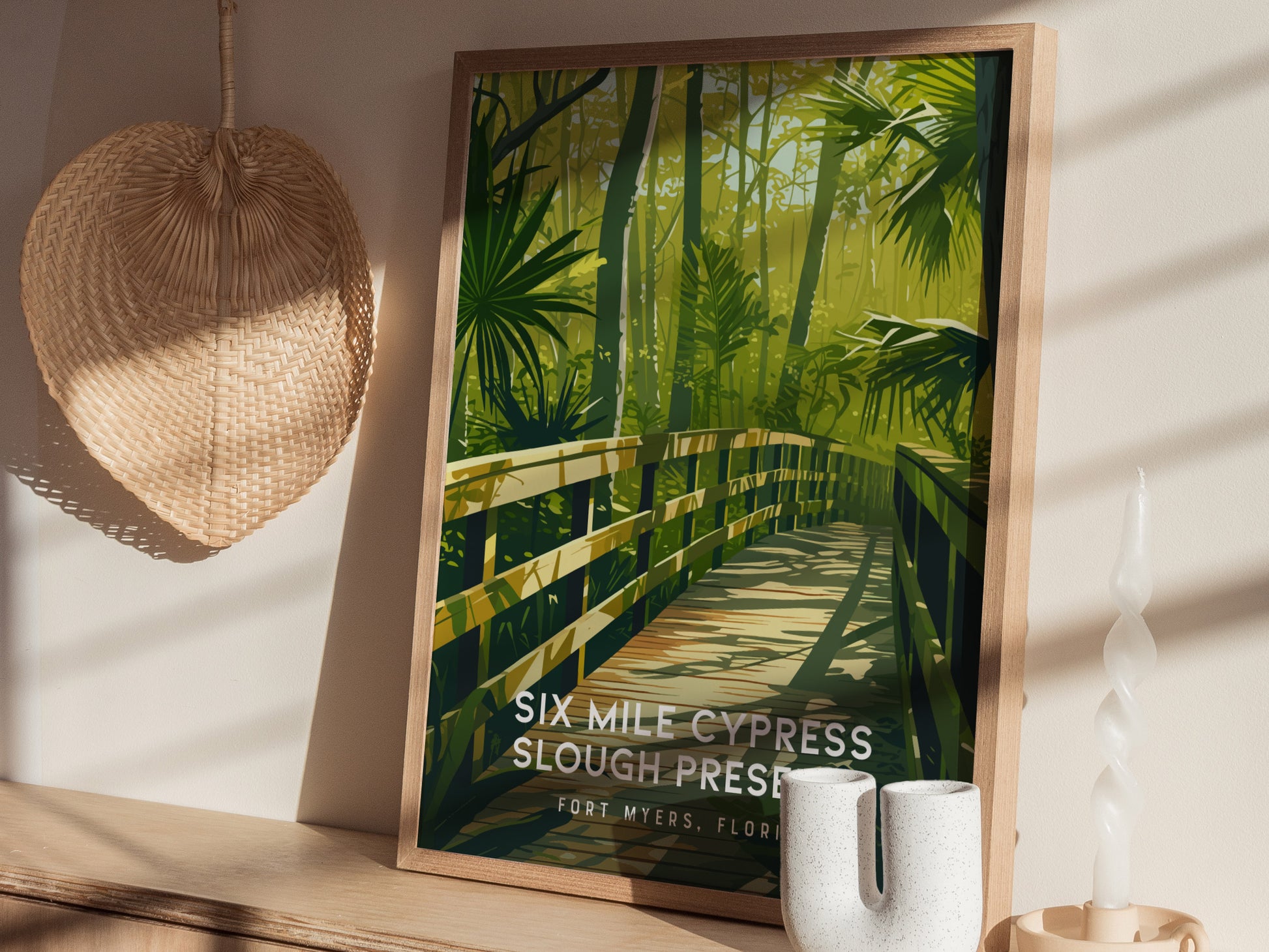 A framed poster of Six Mile Cypress Slough Preserve shows a wooden boardwalk through lush greenery. It is displayed on a shelf with a woven fan and decorative candle.