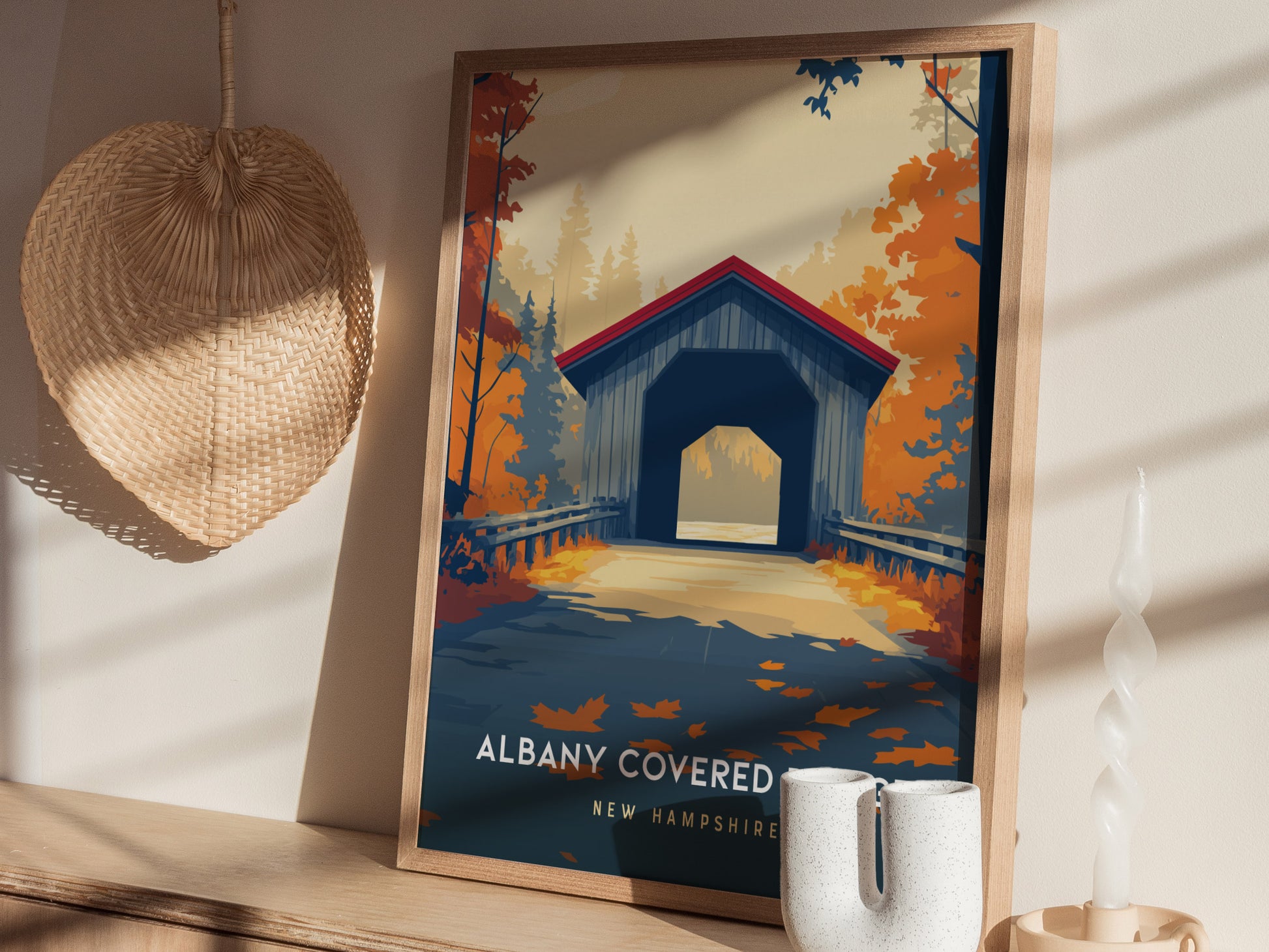 A framed poster of the Albany Covered Bridge in New Hampshire is displayed on a table, featuring vibrant autumn colors with orange and red foliage. The bridge has a blue exterior and a striking red roof, set against a serene background.
