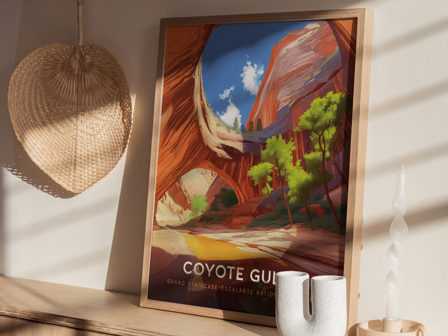 A framed poster of Coyote Gulch with vibrant canyon walls and trees is displayed on a wooden surface, alongside a woven wall decor and a twisted candle.