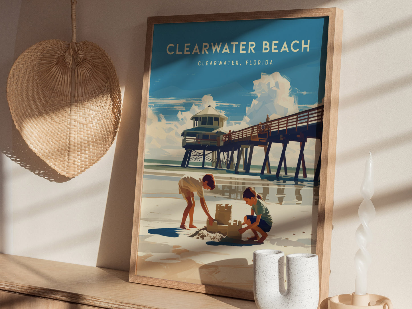 A framed Clearwater Beach poster depicts two children building a sandcastle beside a pier under a blue sky. The scene is complemented by a woven hat and a candle on.