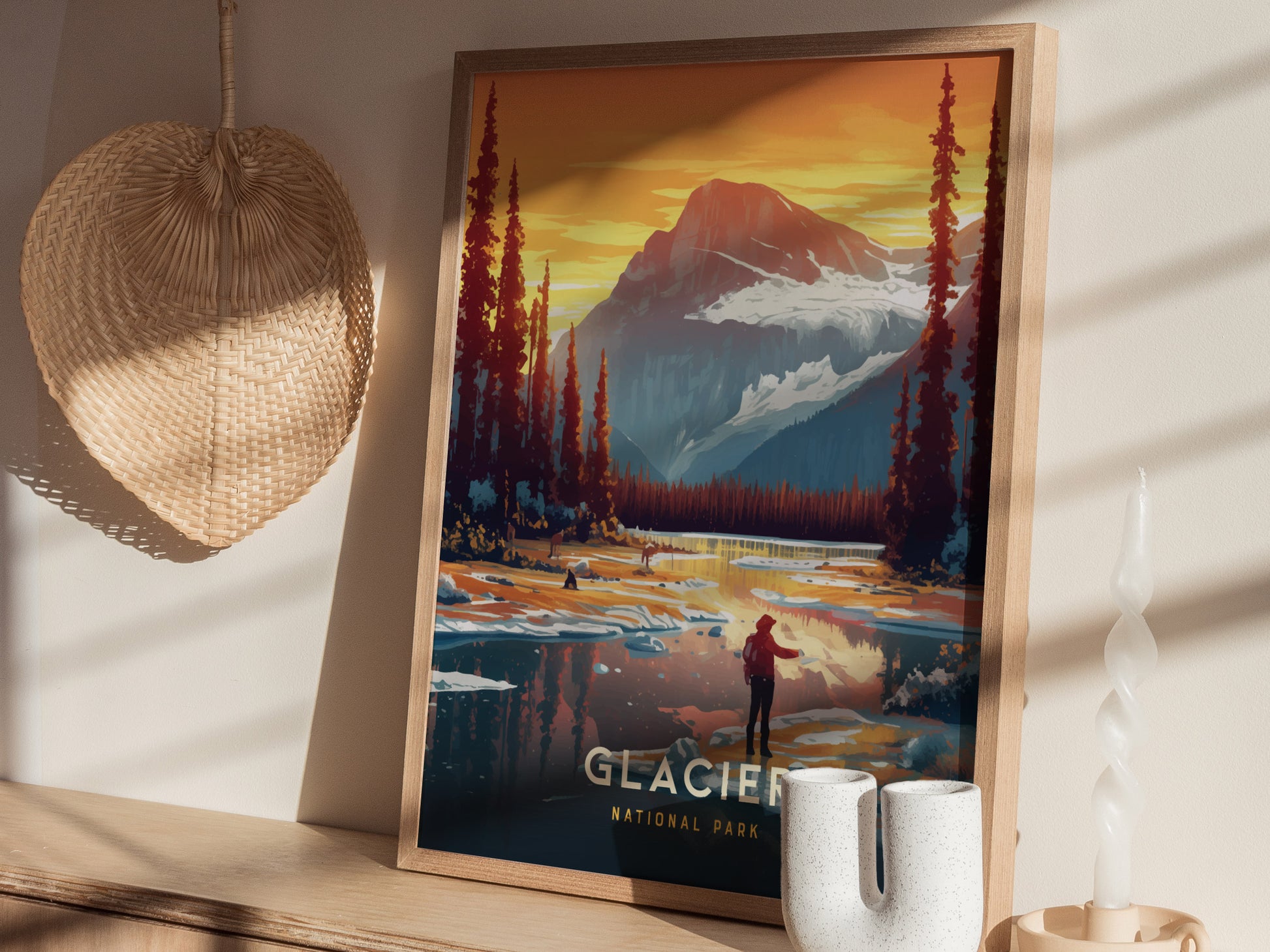 A framed Glacier National Park poster showcases a vibrant mountain landscape with a figure near a reflective lake. It sits on a shelf beside a woven basket and decorative candle.