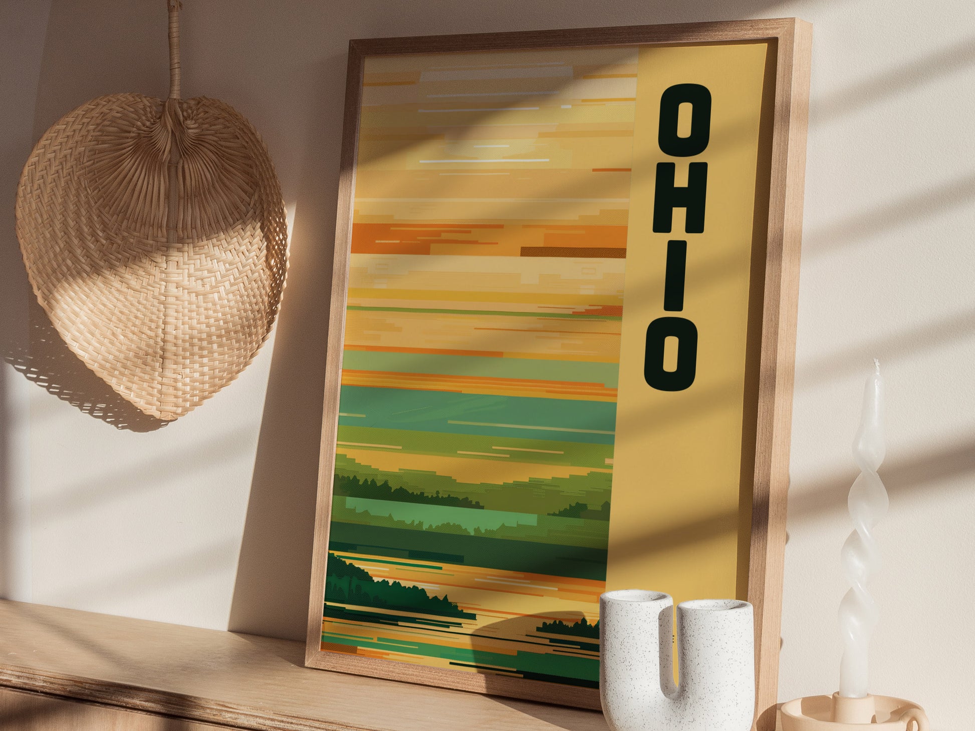 A framed Ohio poster with a minimalist design featuring yellow and green tones is displayed on a wooden shelf. A wicker decoration and a twisted candle are nearby.