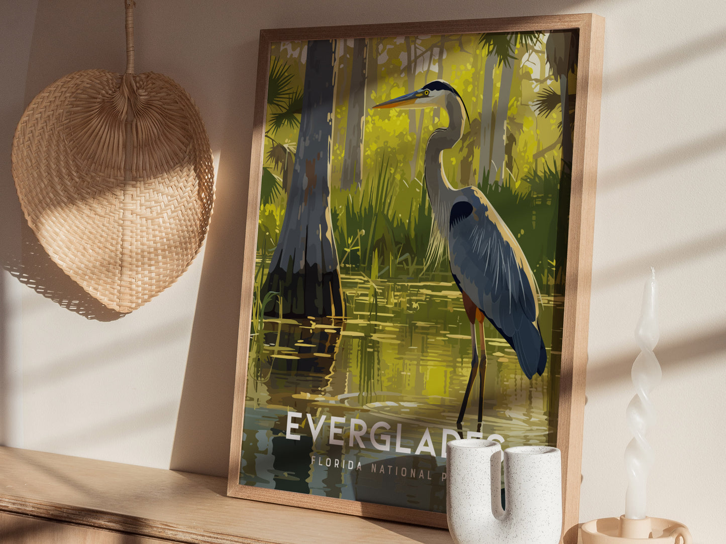 A framed poster of a Great Blue Heron standing in water, surrounded by lush greenery, is displayed on a wooden shelf next to a wicker basket and a twisted candle.