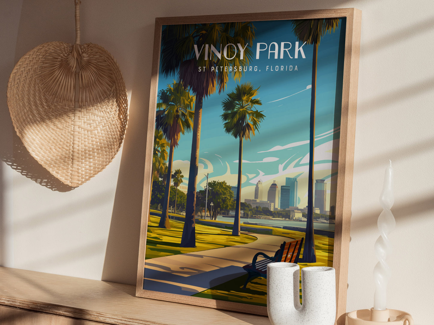A framed Vinoy Park poster rests on a wooden shelf, depicting palm trees and a skyline under a blue sky. Nearby are a woven fan and a twisted candle.