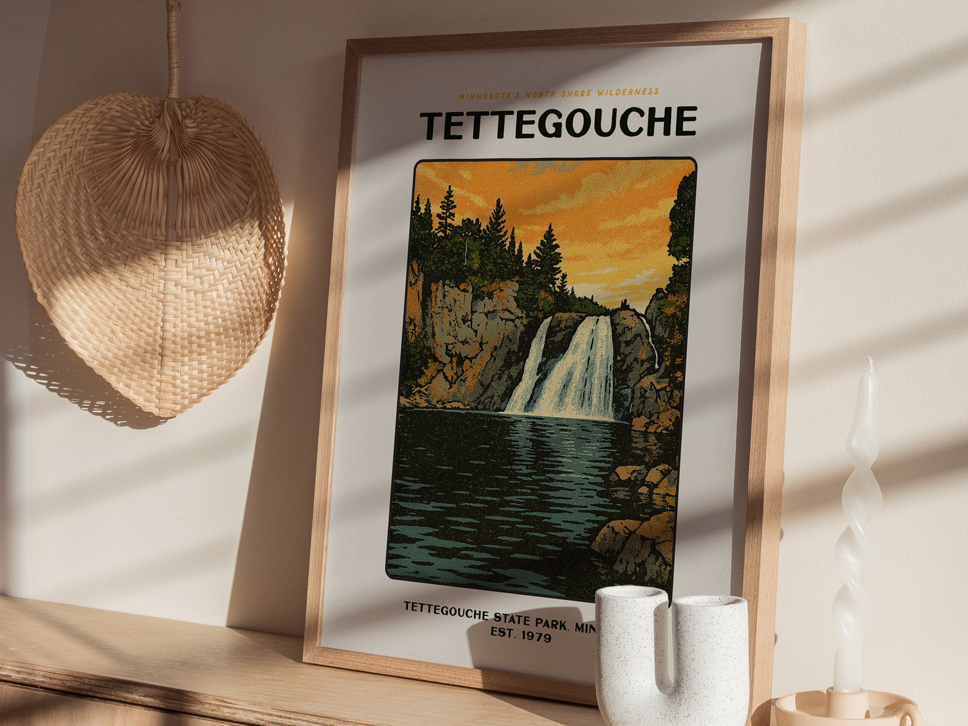 A framed Tettegouche State Park poster featuring a waterfall is displayed on a wooden surface, alongside a woven wall decoration and a unique ceramic vase.