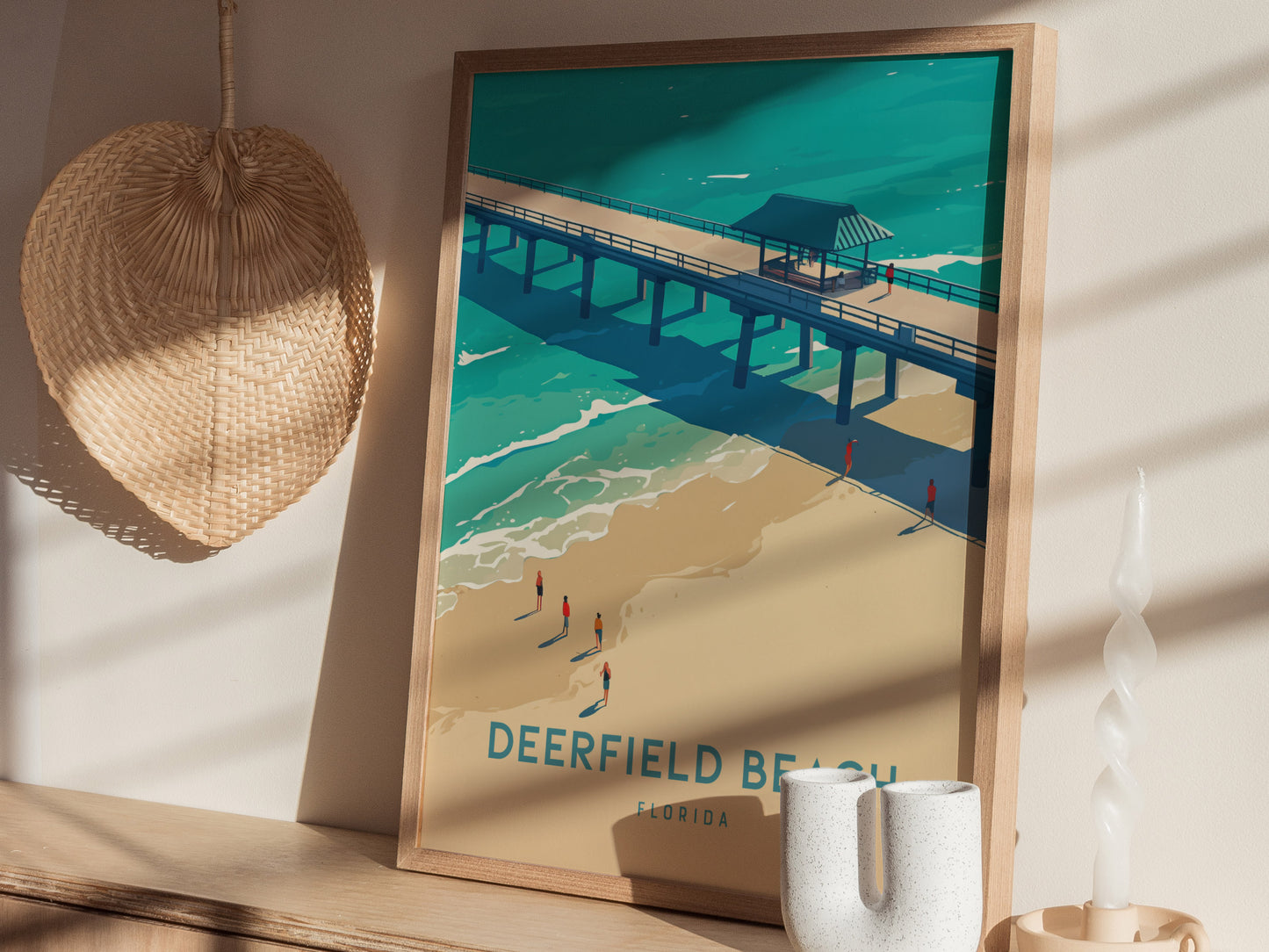 A framed Deerfield Beach poster leans against a wall, depicting a pier over turquoise waters and golden sands. Nearby, a woven hat.